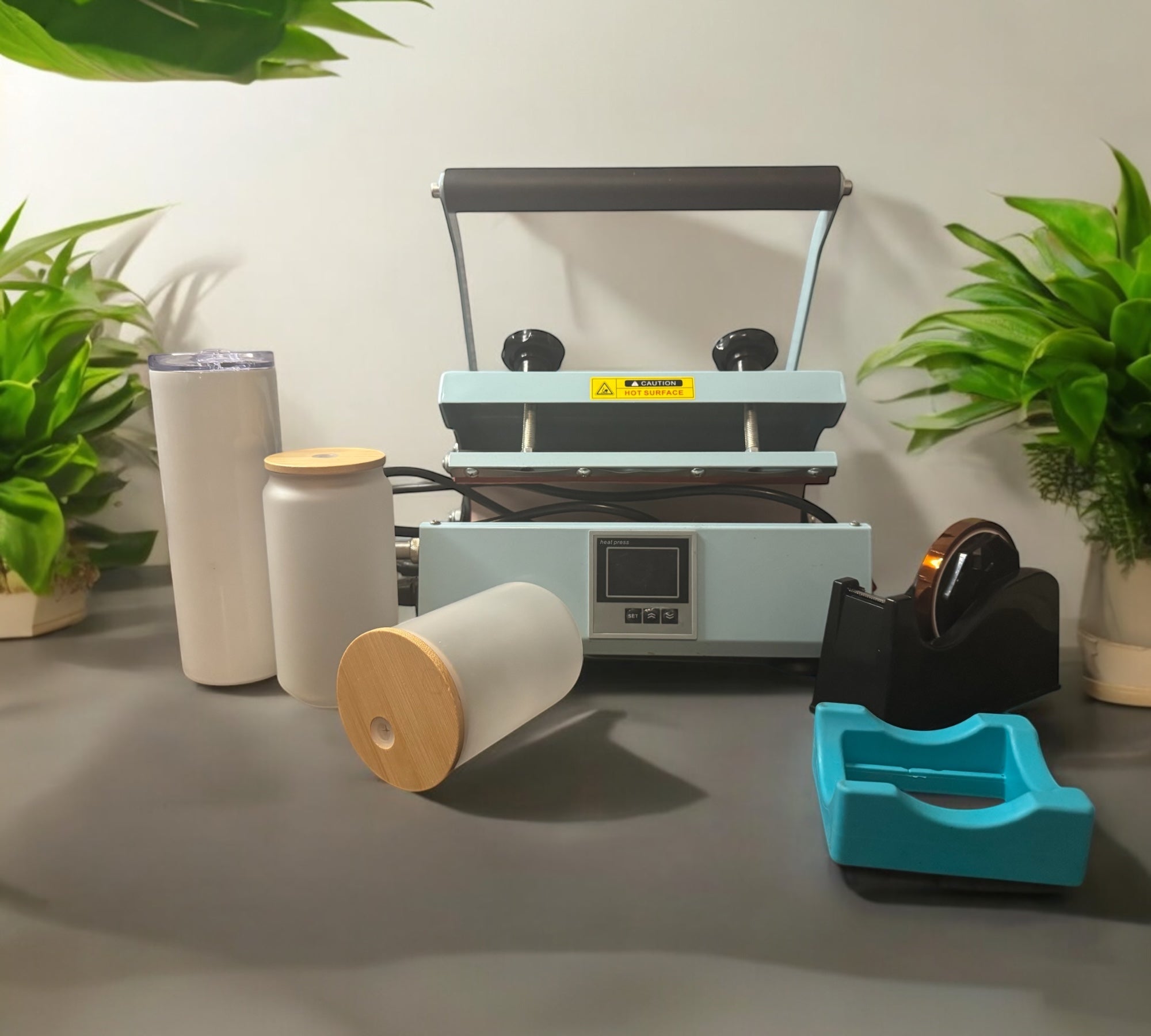 Sublimation machine with blank frosted tumblers and bamboo-lid cups surrounded by green plants.