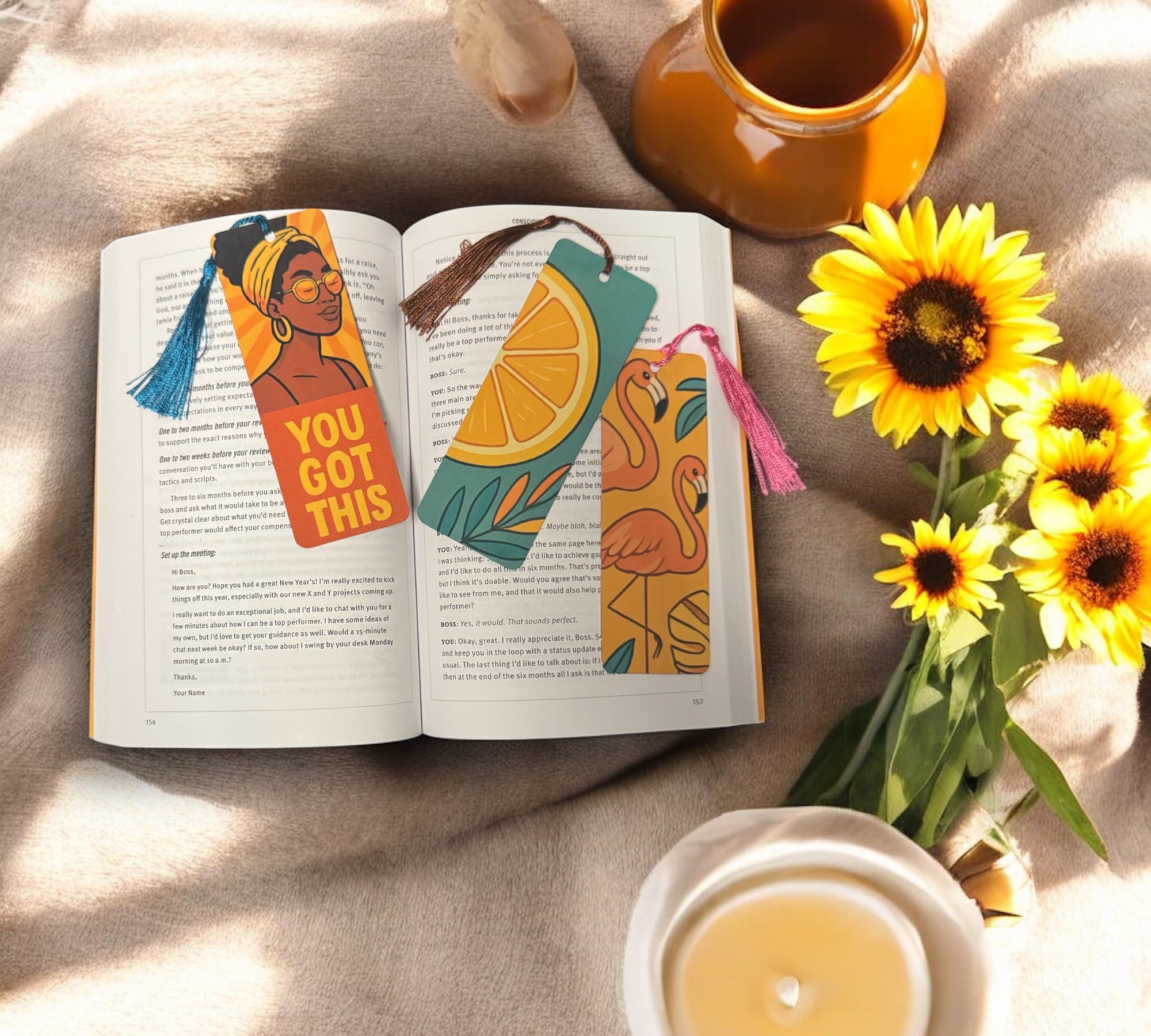 Top-down view of three vibrant summer bookmarks in an open book, styled with sunflowers, a lit candle, and a warm drink on a beige blanket for a cozy seasonal vibe.